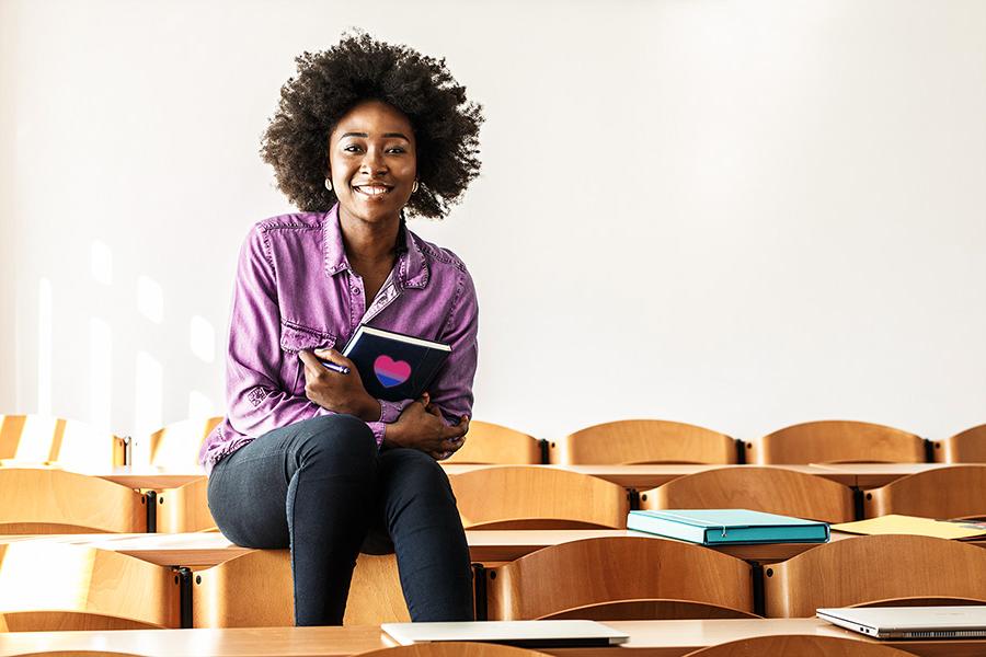 一个非裔美国学生坐在大学教室里，手里拿着一个笔记本，上面有心形的双性恋旗帜